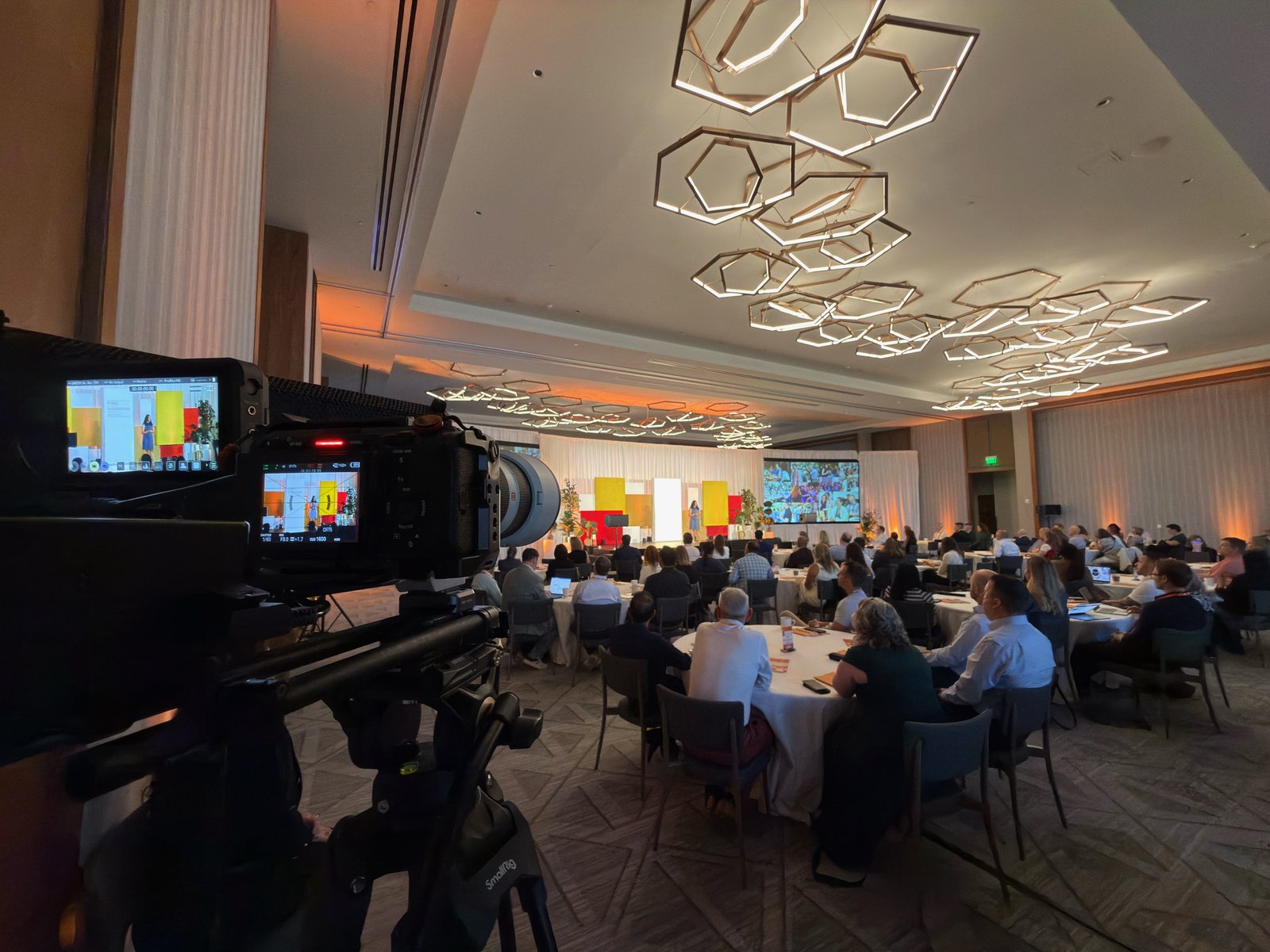 A cinema camera at the back of a full conference room, speaker on stage beyond