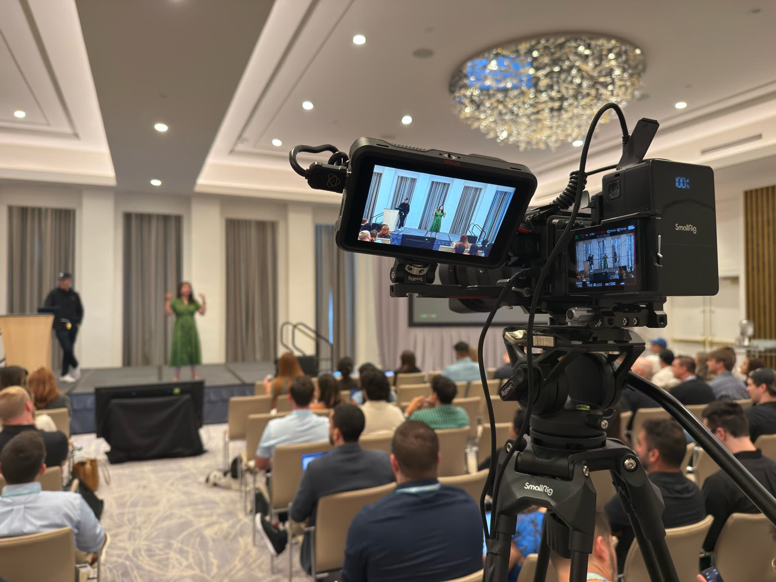 A camera and monitor framing a speaker on a ballroom main stage, the audience seated beyond